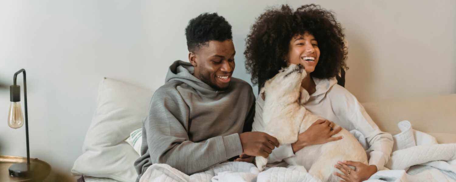 Couple sitting on a bed with a dog, smiling and holding the dog.
